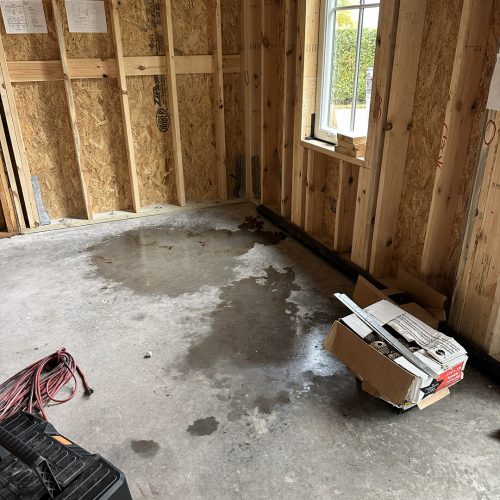 1. Interior view of a newly constructed home under renovation showing exposed wooden framing, a concrete floor with water stains, and a window providing natural light.