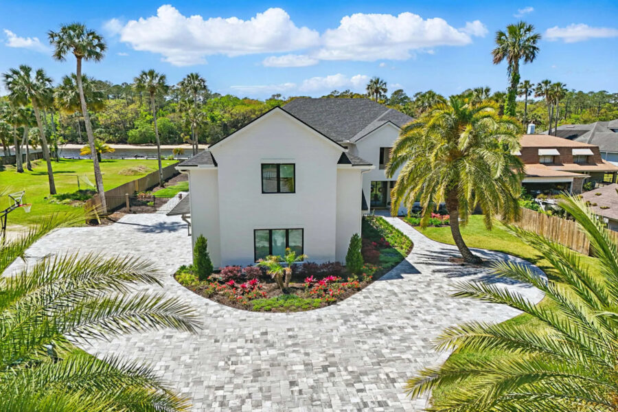 Modern white two-story home with a paved driveway, surrounded by lush tropical landscaping and palm trees.