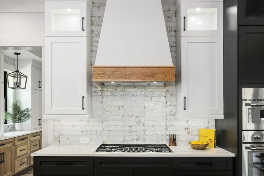 Contemporary kitchen featuring white cabinetry, black lower cabinets, a white brick backsplash, and a wooden range hood, creating a sleek and stylish cooking space.