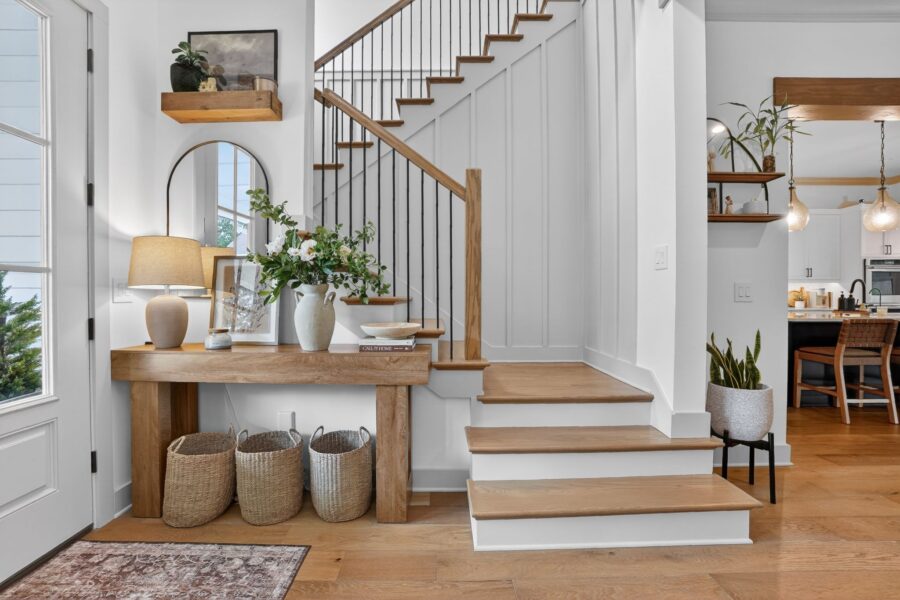 A bright, welcoming entryway featuring a staircase with wooden steps and black metal railing, a wooden console table with decorative items, and a cozy rug, creating a stylish and inviting home entranc.