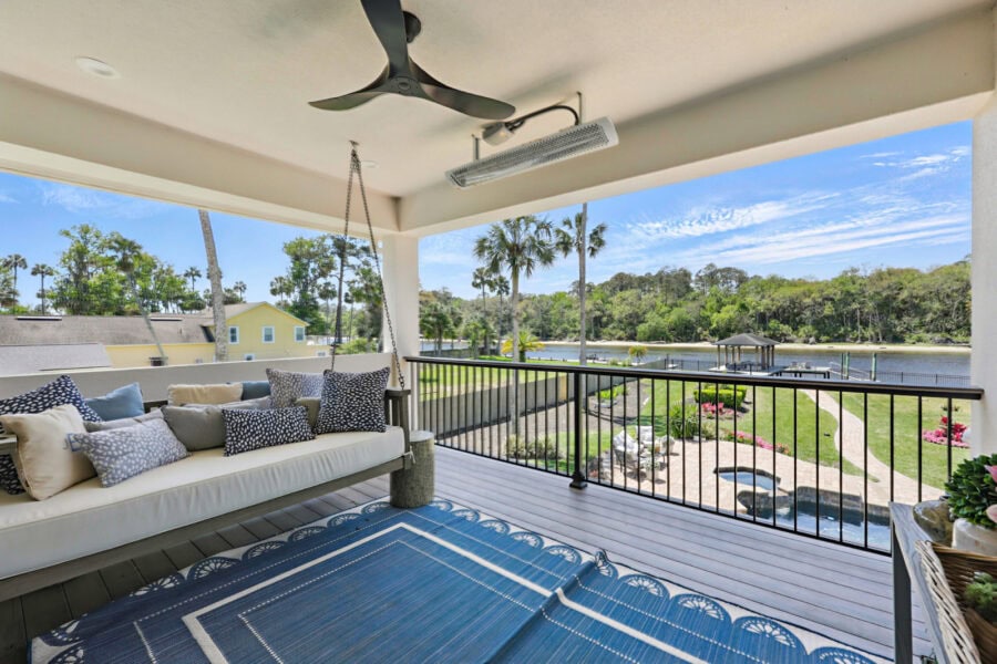 Bright covered porch with a view of a lush landscaped yard, a pond, and a dock, featuring outdoor seating, a ceiling fan, and decorative cushions for a relaxing outdoor living space.