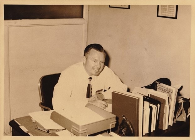 Man in white dress shirt and tie sitting at desk filled with papers and office supplies, vintage office setting, black and white photo, early to mid-20th century.