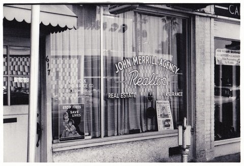 John merrill agency real estate office storefront with signage and window displays, black and white.